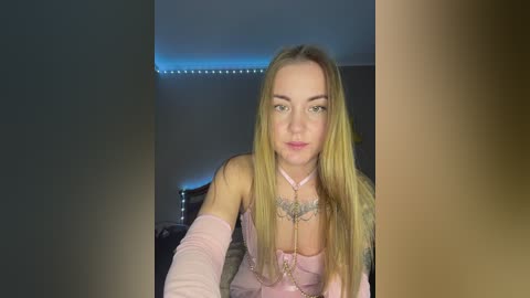 Media: Video of a blonde Caucasian woman in lingerie with a choker, tattoos, and long hair, sitting in a dimly lit bedroom with a blue LED strip light.