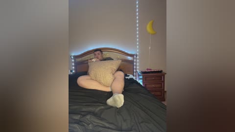 Media: Video of a light-skinned woman with short dark hair, wearing a beige knit sweater and white socks, sitting on a bed with a dark comforter, surrounded by string lights and a yellow crescent moon decoration on a wooden nightstand.