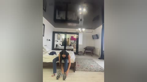 Media: Video of a woman bending down to tie her shoes in a modern, minimalist bedroom with a mirror, chandelier, and patterned rug.