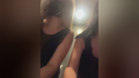 Media: A blurry video of a woman in a dark, possibly nightclub setting. She has long, dark hair and is wearing a black sleeveless top. The image is dimly lit with a bright light source in the background, creating a hazy effect.