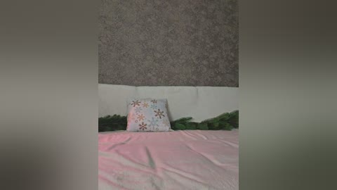 Media: A video of a bed with a white pillow featuring floral patterns, against a grey carpeted wall and a white wall. The bed is covered with pink sheets and green ferns.