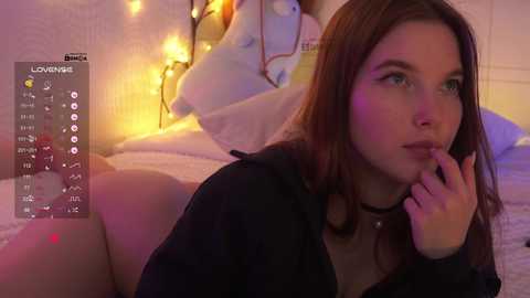 Video of a young woman with fair skin, long brown hair, wearing a black choker and black top, lying on a bed with a white duvet, surrounded by string lights, and a digital clock overlay.