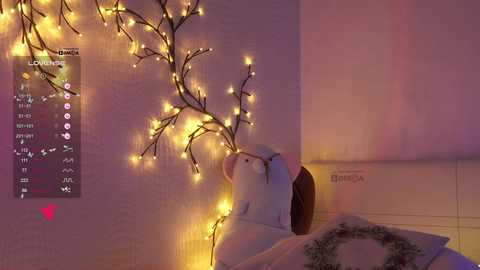 Video of a woman with fair skin, long brown hair, and a white shirt, lying on a bed. She holds a tree-shaped string of warm yellow lights against her face, casting a soft glow. The background features a light pink wall and a white headboard.