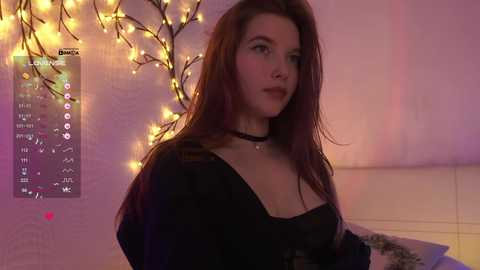 Video of a young woman with long, straight brown hair, wearing a black top and choker, standing in a softly lit room with warm lighting and a decorative calendar.