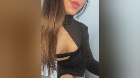 Media: Video of an Asian woman with long, straight brown hair, wearing a black, high-neck crop top with a large cutout revealing her left breast. The background features a brown wall on the left and a grey wall on the right.