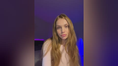 A video of a young woman with long, wavy, light brown hair and fair skin, wearing a white top, against a dark, purple-lit background. She has a neutral expression and stands centrally.