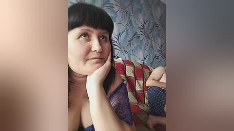 Video of a fair-skinned woman with short black hair and bangs, wearing a purple lace bra, resting her chin on her hand, against a colorful, floral-patterned wallpaper background.