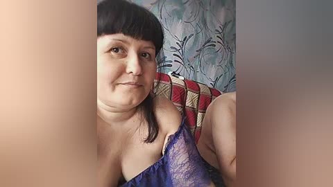 Video of a plus-size woman with straight black hair and bangs, wearing a purple lace bra, sitting on a red and white patterned chair against a floral-patterned wall.