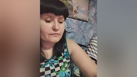 Video of a young woman with long black hair and fair skin, wearing a sleeveless dress with a black and white checkered pattern and blue floral print, sitting indoors with a blue and green floral wallpaper in the background.