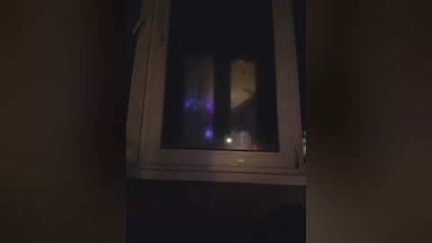 A dimly lit video captures the interior of a dark room with a single window. The window frame is white, and the glass is slightly reflective, showing faint, indistinct light sources outside. The room appears sparsely furnished.