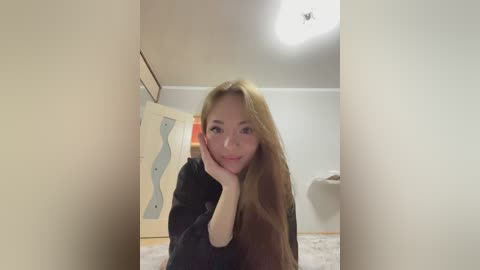 Media: Video of a young Asian woman with long, straight, blonde hair, wearing a black sweater, leaning against a beige wall in a minimalist room with a light carpet and white walls.