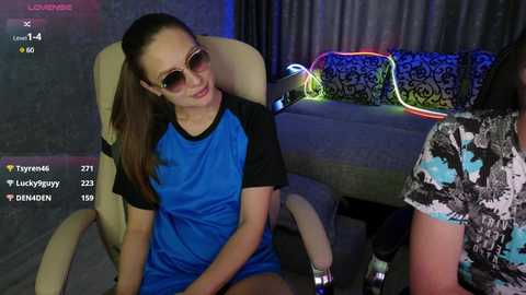 Video of a young woman in a blue and black t-shirt, sunglasses, and a ponytail, seated on a beige chair in a dimly lit room with a gray couch and colorful pillows.