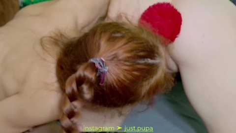 Media: A video shows a close-up of a person with light skin and long brown hair, lying on a bed with a red, fuzzy butt plug inserted.