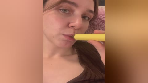 Media: A close-up video of a light-skinned woman with straight brown hair, wearing a black top, sucking on a yellow vibrator. The background features a pink fluffy blanket.