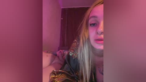Video of a young woman with long blonde hair, multiple piercings, and tattoos, wearing a colorful floral shirt. The background features dim lighting and a closed door, giving a cozy, intimate indoor setting.
