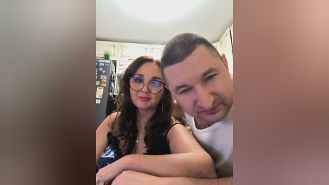 Media: Video of a smiling, fair-skinned couple with medium builds and short hair. Woman with glasses, wearing a black top, embraces a man in a white shirt. Background features a fridge and kitchen counters.