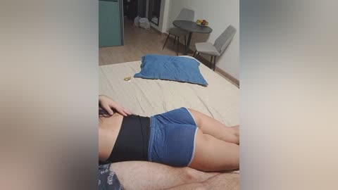 Video of a woman lying on a bed, wearing blue shorts and a black sports bra, with a gray table and chairs in the background.