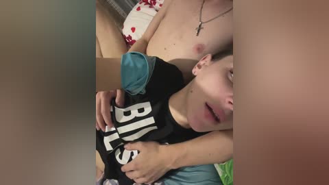 Media: Video of a shirtless man with fair skin and small nipples cradling a young boy with dark hair, wearing a black shirt with \"BIG\" in white letters. They are in a bed with red and white patterned pillows.