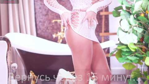 Media: Video of a slender woman in a sheer, white, fishnet dress, highlighting her toned legs, standing in a luxurious, vintage-style room with a clawfoot tub, greenery, and ornate decor.