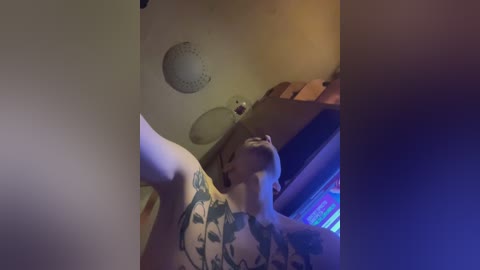 Media: A dimly-lit video shows a tattooed, shirtless man with short hair and glasses, lying on a bed with a white ceiling fan and colorful neon lights.