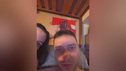 Media: Video of a young man with short dark hair lying on a bed, partially obscured by a pillow, with a modern abstract painting and wooden headboard in the background.