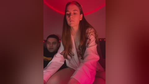 Media: A video of a young woman with long brown hair, wearing a white robe, sitting on a bed with a man behind her. The room is dimly lit with red lighting.