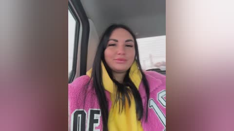 A video of a young woman with long black hair, wearing a pink and yellow blanket, sitting in a car with blurred background.
