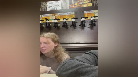 Media: Video of a blonde woman in a beige sweater, seated at a table, with a dark-haired man's legs visible under the table. Behind them, a row of coffee machine spouts and a menu board with \"Buddy\" branding.