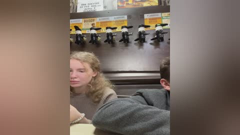 Media: Video of a woman with curly hair in a beige sweater, sitting at a table with a man in a gray jacket, in a diner with a beer tap and menu signs.