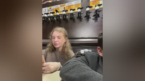 Media: Video of a young woman with long, curly blonde hair, wearing a beige sweater, sitting at a table in a dimly lit room with a dark wooden wall and microphone stands.