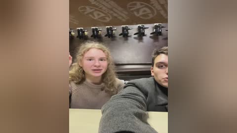 Media: Video of two young people with curly hair in a dimly lit room. The girl in a beige sweater is smiling, while the boy in a gray hoodie looks serious. A dark wooden wall with hooks and a decorative pattern is visible in the background.