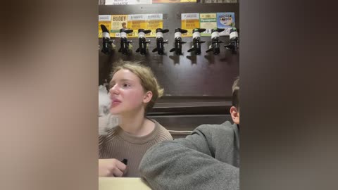 Media: Video of a young woman with light skin and short blonde hair, wearing a beige sweater, sitting at a table in a dimly lit restaurant. Behind her, mounted black-and-white penguin figures and colorful menu signs hang on a dark wall.