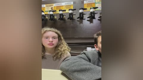 Media: Video of two people in a bathroom with a beige wall and multiple black faucets. A young woman with curly hair, wearing a knit sweater, stands beside a man in a gray jacket, both looking away.