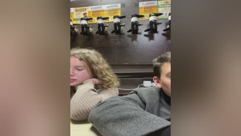 Media: Video of two people in a dimly lit, beige-colored room; one person, with curly blonde hair, is asleep on a beige couch, while the other, with short dark hair, is sitting nearby, looking at the person asleep.