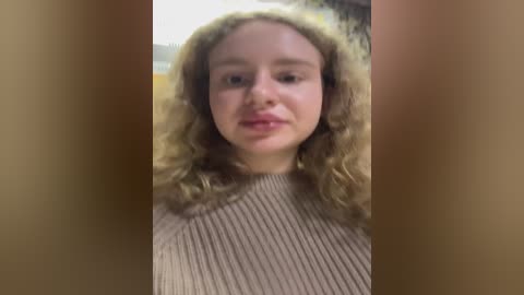 Media: Video of a young woman with curly blonde hair, fair skin, and wearing a beige ribbed sweater. She has a neutral expression and is partially obscured by a blurry background.
