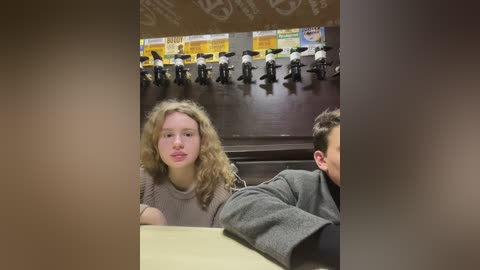 Media: Video of a young woman with curly blonde hair in a grey sweater sitting in a classroom with a projector screen displaying a cartoon image.