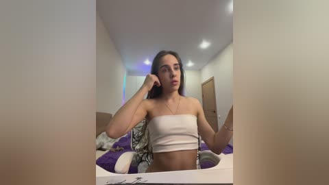 Media: Video of a young woman with long, dark hair, wearing a white strapless crop top, sitting on a bed with purple and white bedding, talking on a phone, in a modern, minimalist bedroom.