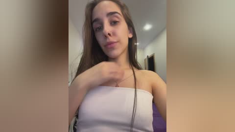 Media: Video of a young woman with fair skin, long dark hair, and brown eyes, wearing a white strapless top. She is indoors, looking at the camera with a neutral expression.