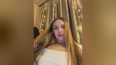 Video of a young Caucasian woman with long, straight blonde hair, light skin, and a slender physique, wearing a white off-shoulder top. She stands in front of a gold and cream floral-patterned curtain in a softly lit room.