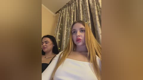 Video of two young women, one with long blonde braids and red lipstick, standing close, in a dimly lit room with beige walls and gold-patterned curtains.