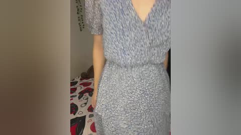 Media: Video of a woman wearing a blue and white polka-dotted dress, standing indoors with a floral-patterned bedspread visible in the background.