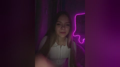 Video of a young woman with light brown hair, wearing a white top, smiling in a dimly lit room with purple neon lighting, creating a moody atmosphere.