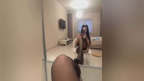 Media: A video of a young woman with long dark hair, posing in a mirror, wearing sheer black stockings and a bra, in a minimalist bedroom with beige walls and a bed.