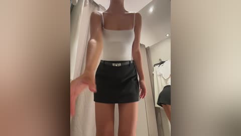 Media: Video of a slim, light-skinned woman with straight brown hair, wearing a tight white tank top and short black skirt, standing in a beige-walled dressing room, with clothes hanging nearby.