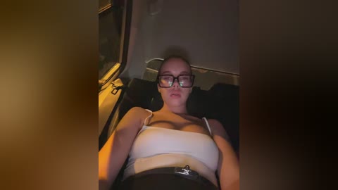 Media: Video of a young woman with fair skin, wearing glasses, a white tank top, and black skirt, reclined in a car at night, dim lighting, slightly out of focus.