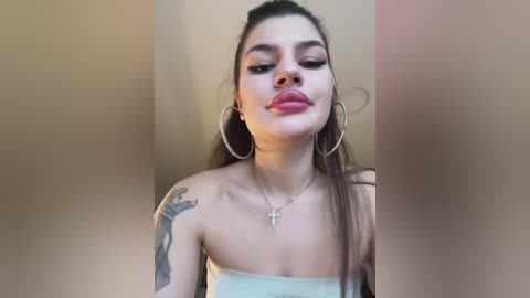 Media: Video of a young Latina woman with long dark hair, wearing large silver hoop earrings and a white strapless top, posing with a pouty face and a tattoo on her left shoulder.