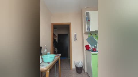 Media: A video of a small, cluttered kitchen with beige walls and wooden furniture. A person in a dark jacket stands in an adjacent room, while a green bowl sits on a wooden table.