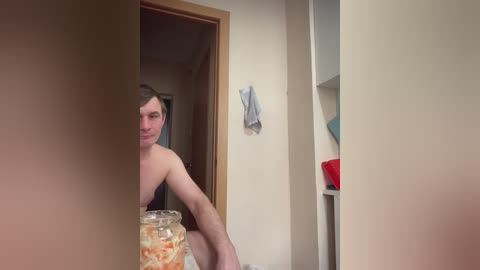 Media: Video of a shirtless man with short brown hair, sitting on a bed with a jar of pickles in the foreground, in a small, cluttered room with beige walls and a wooden door.
