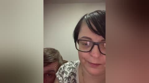 Media: Video of a middle-aged woman with short black hair, glasses, and a patterned shirt, smiling, with an older man with gray hair in the background.