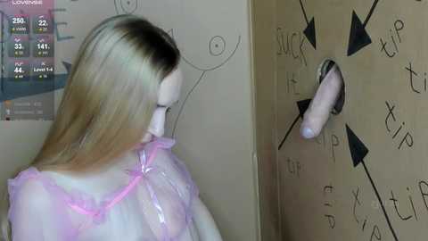 Media: Video of a blonde woman in a pink see-through dress, facing a dartboard with a dildo attached to it, in a dimly lit room.